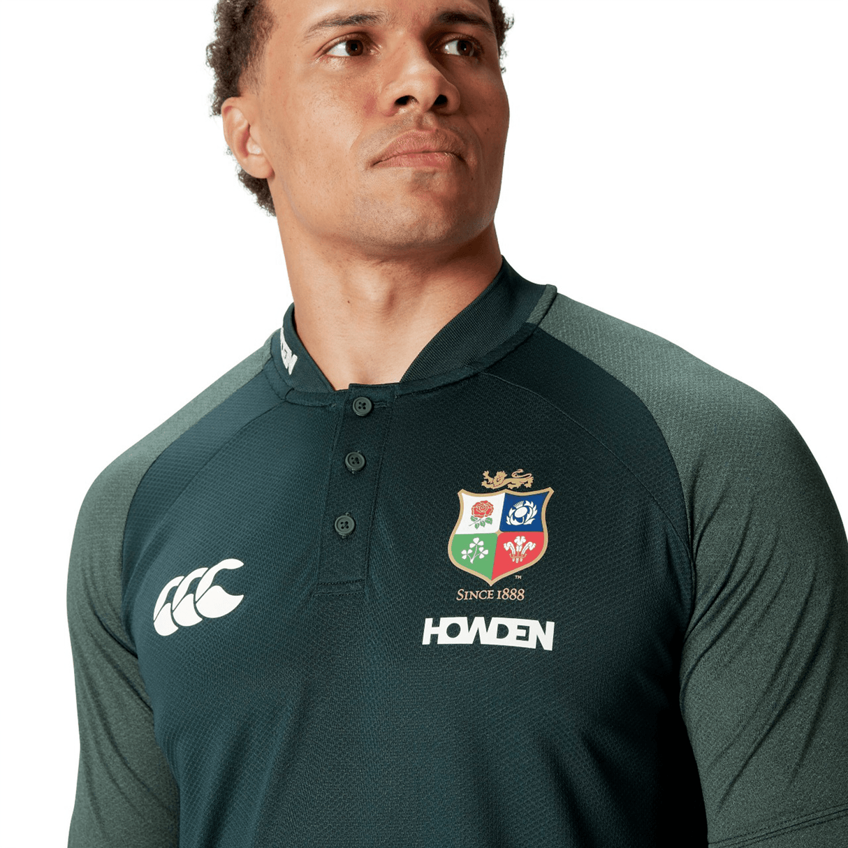 A person wearing the British & Irish Lions 25 Poly Polo by Canterbury, featuring a dark green color with a multicolored crest and brand logos on the front. Made from breathable, wicking fabric, it promises maximum comfort and quick drying while keeping your style effortless.