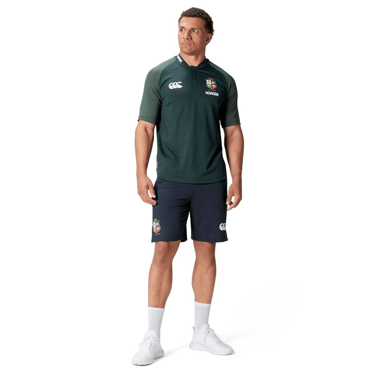 A man stands wearing the British & Irish Lions 25 Poly Polo by Canterbury, a dark green and black sports uniform made of breathable wicking fabric, adorned with logos. He pairs it with white socks and sneakers against a white background.