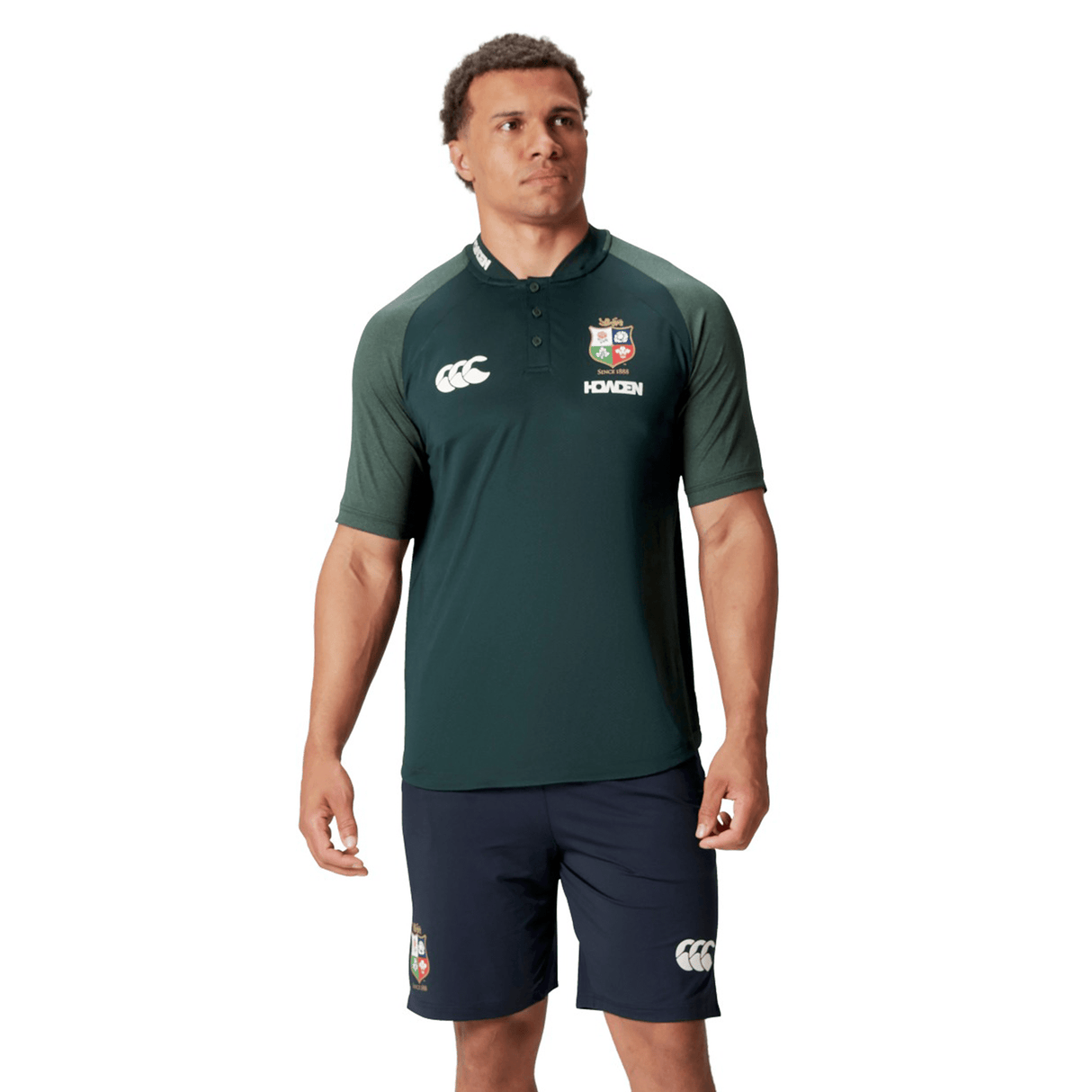 A person wearing the British & Irish Lions 25 Poly Polo by Canterbury, a green and black sports shirt featuring a logo and made from breathable wicking fabric, paired with navy shorts, stands against a white background.
