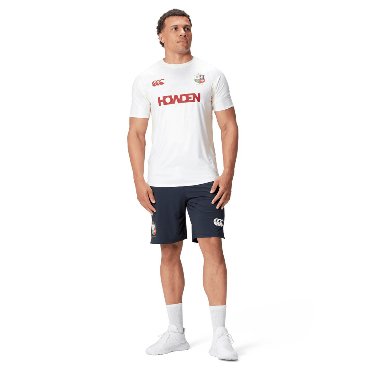 A man is wearing a white sports t-shirt with "Howden" printed in red and British & Irish Lions 2025 Training Shorts by Canterbury, standing against a white background.
