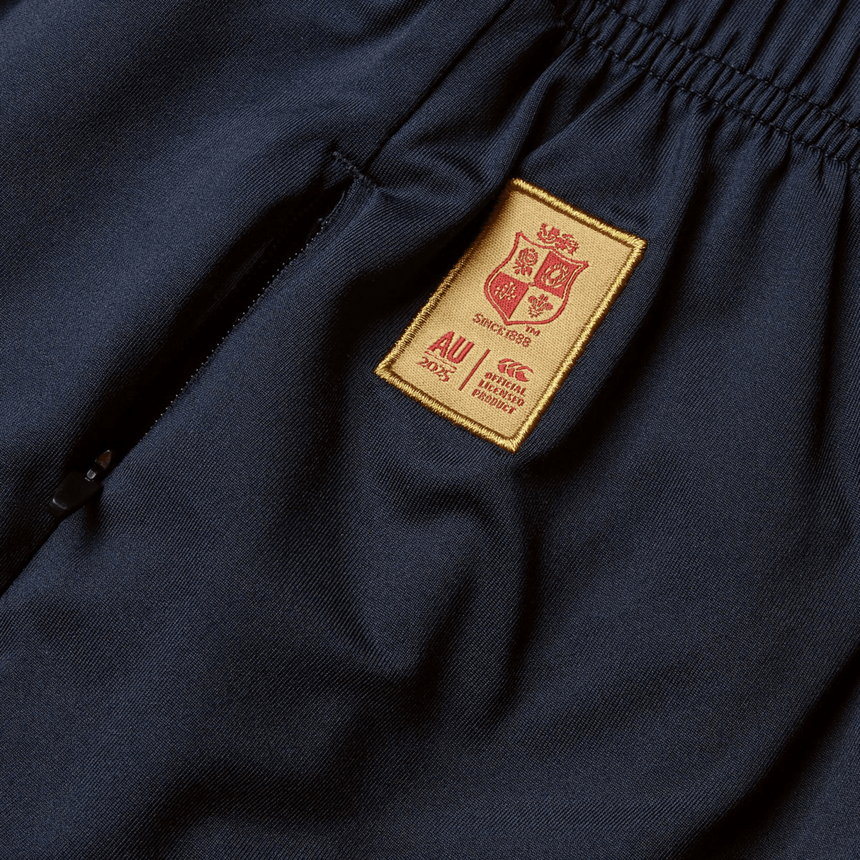 Close-up of navy blue pants by Canterbury, featuring an embroidered gold patch with a coat of arms and text details, reminiscent of the iconic British & Irish Lions 2025 Training Short.