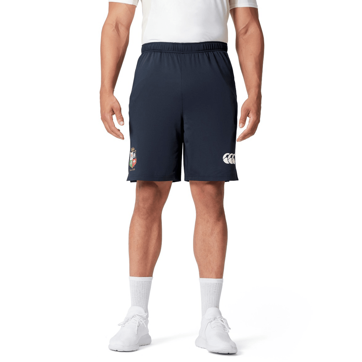 A person wearing a white shirt, British & Irish Lions 2025 Training Short by Canterbury in a dark color with the logo visible, white socks, and white shoes stands against a white background.