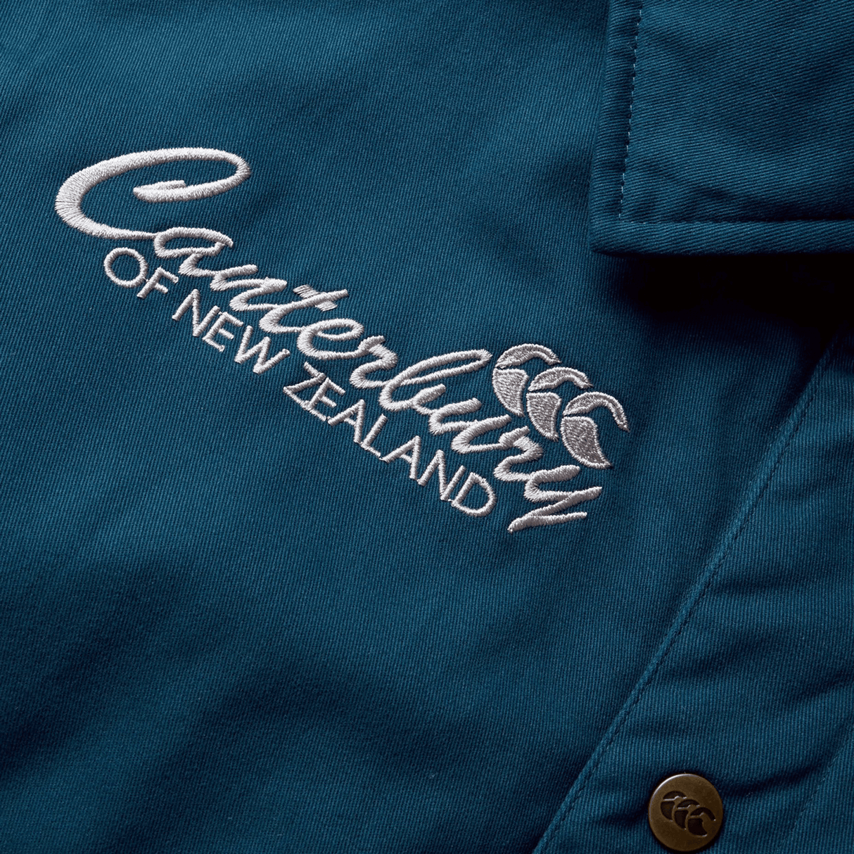 A close-up of the Canterbury logo, embroidered in white on the blue fabric of the British & Irish Lions 2025 Heritage Quilted Jacket by Canterbury, with a metal button visible—an iconic symbol celebrating the legacy of the Lions.