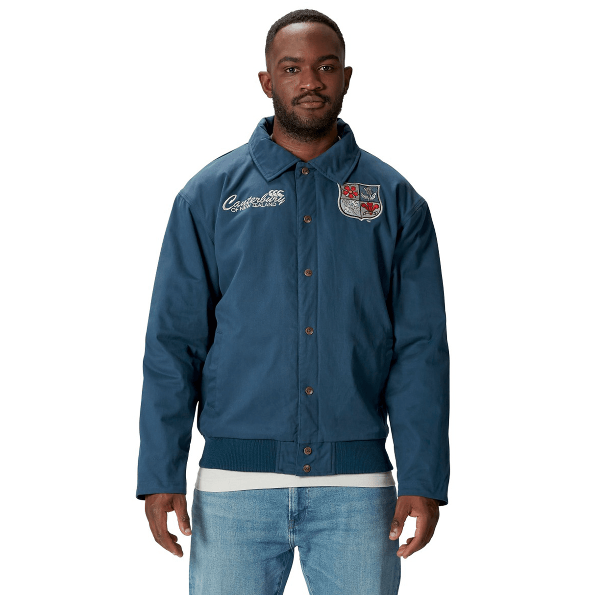A man wearing a blue British & Irish Lions 2025 Heritage Quilted Jacket by Canterbury, embellished with patches, stands against a white background, showcasing the timeless style synonymous with Canterbury and the British & Irish Lions.