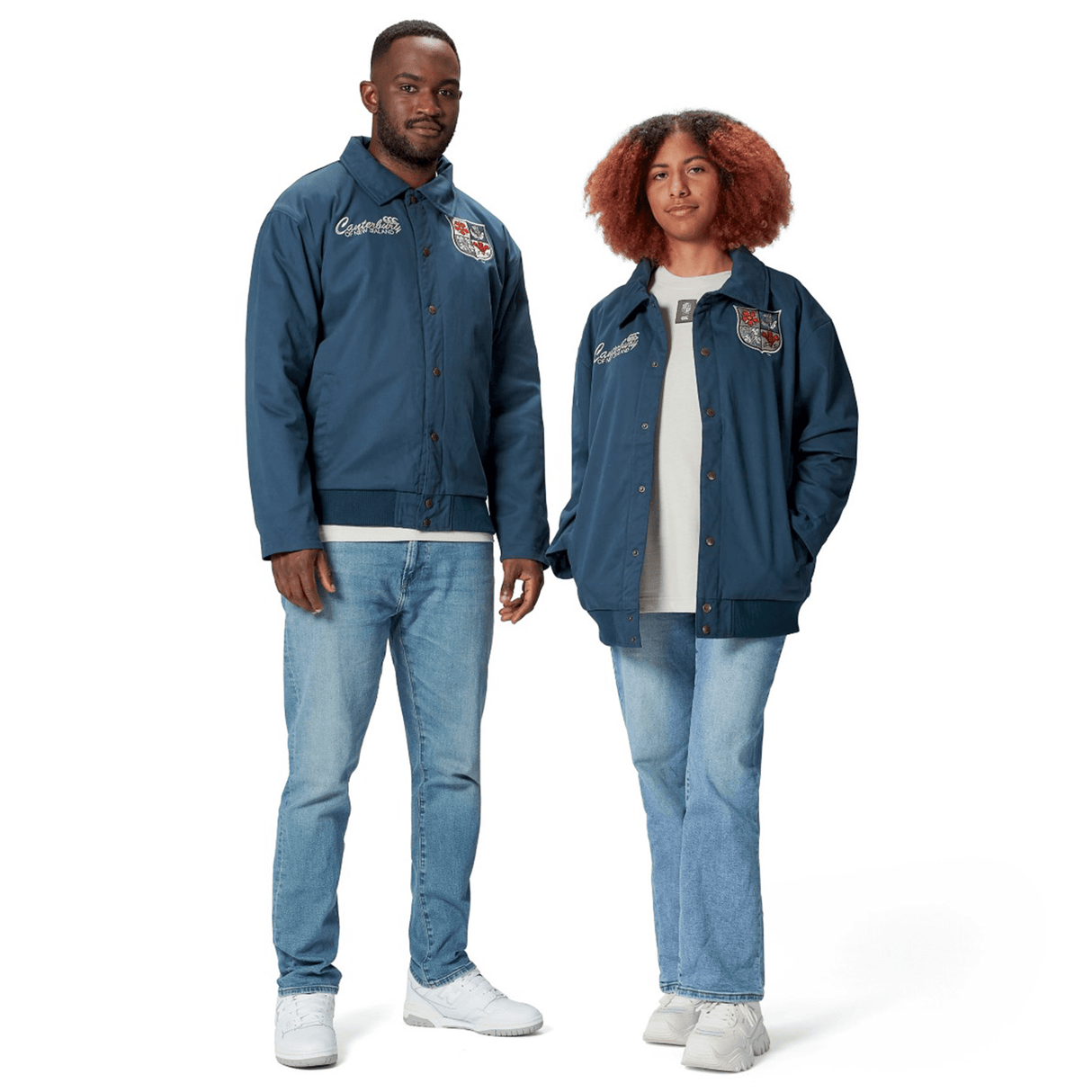 Dressed in matching British & Irish Lions 2025 Heritage Quilted Jackets by Canterbury, adorned with embroidered patches that celebrate the team's legacy, two individuals pose elegantly. They complement their outfits with jeans and white shoes against a simple white background.
