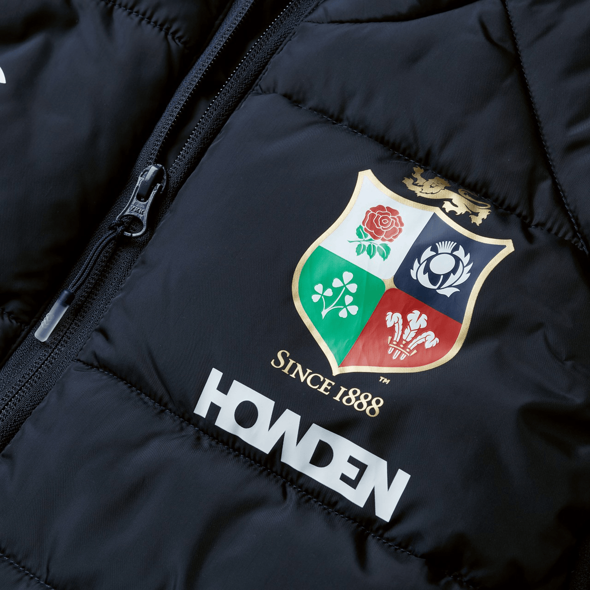 Close-up of a black British & Irish Lions 2025 Microlight Gilet by Canterbury, crafted from recycled polyester. It showcases a colorful embroidered crest with floral motifs and the text "Since 1888," with the word "HOWDEN" printed below the crest.