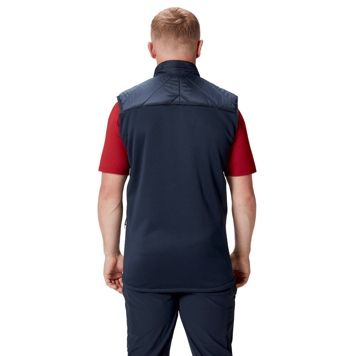 A person with short blonde hair is standing, facing away, wearing a British & Irish Lions 2025 Microlight Gilet by Canterbury made from recycled polyester over a red short-sleeve shirt and navy pants.