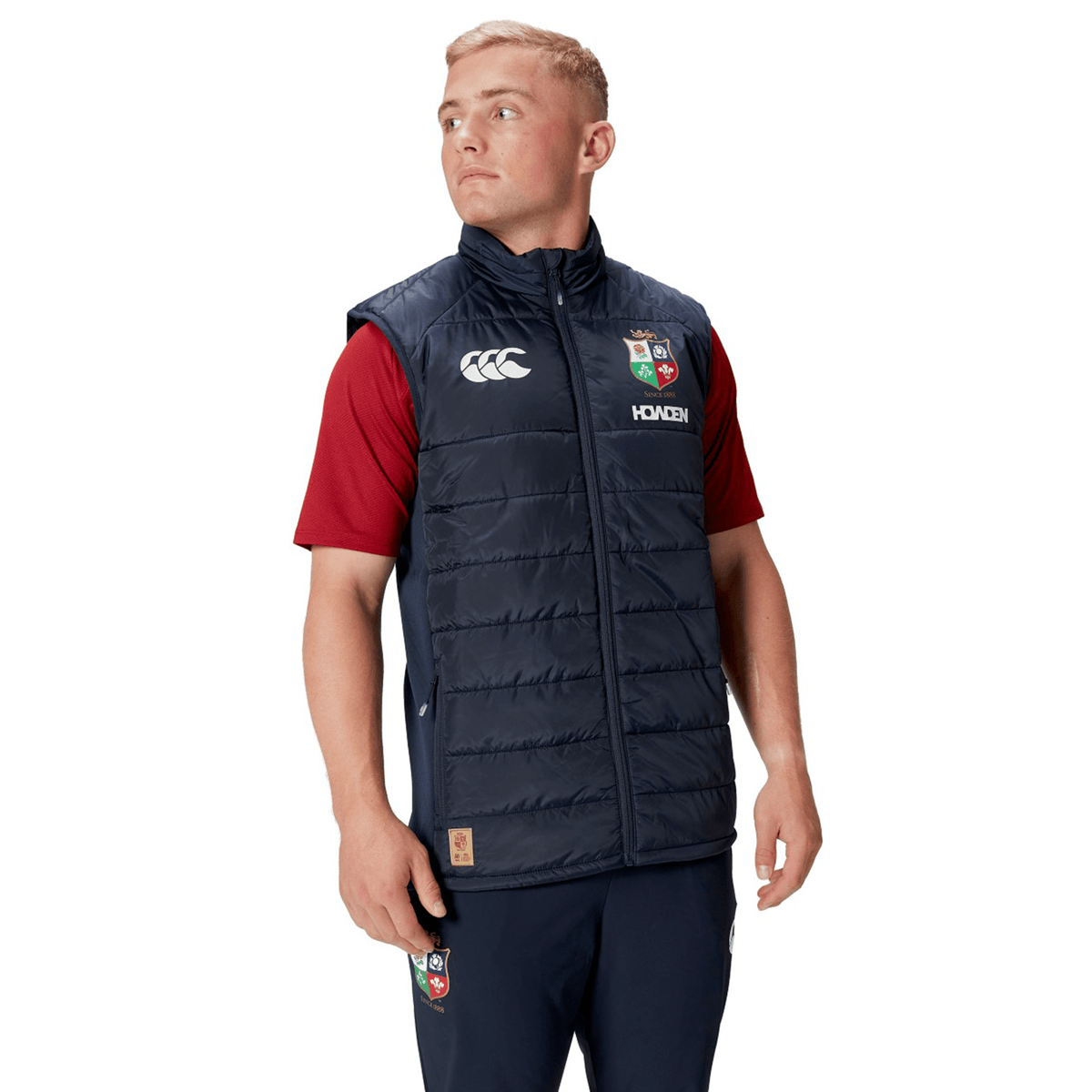 A person wearing a British & Irish Lions 2025 Microlight Gilet by Canterbury in navy blue, made from recycled polyester, over a red shirt and navy pants, looks to the side.