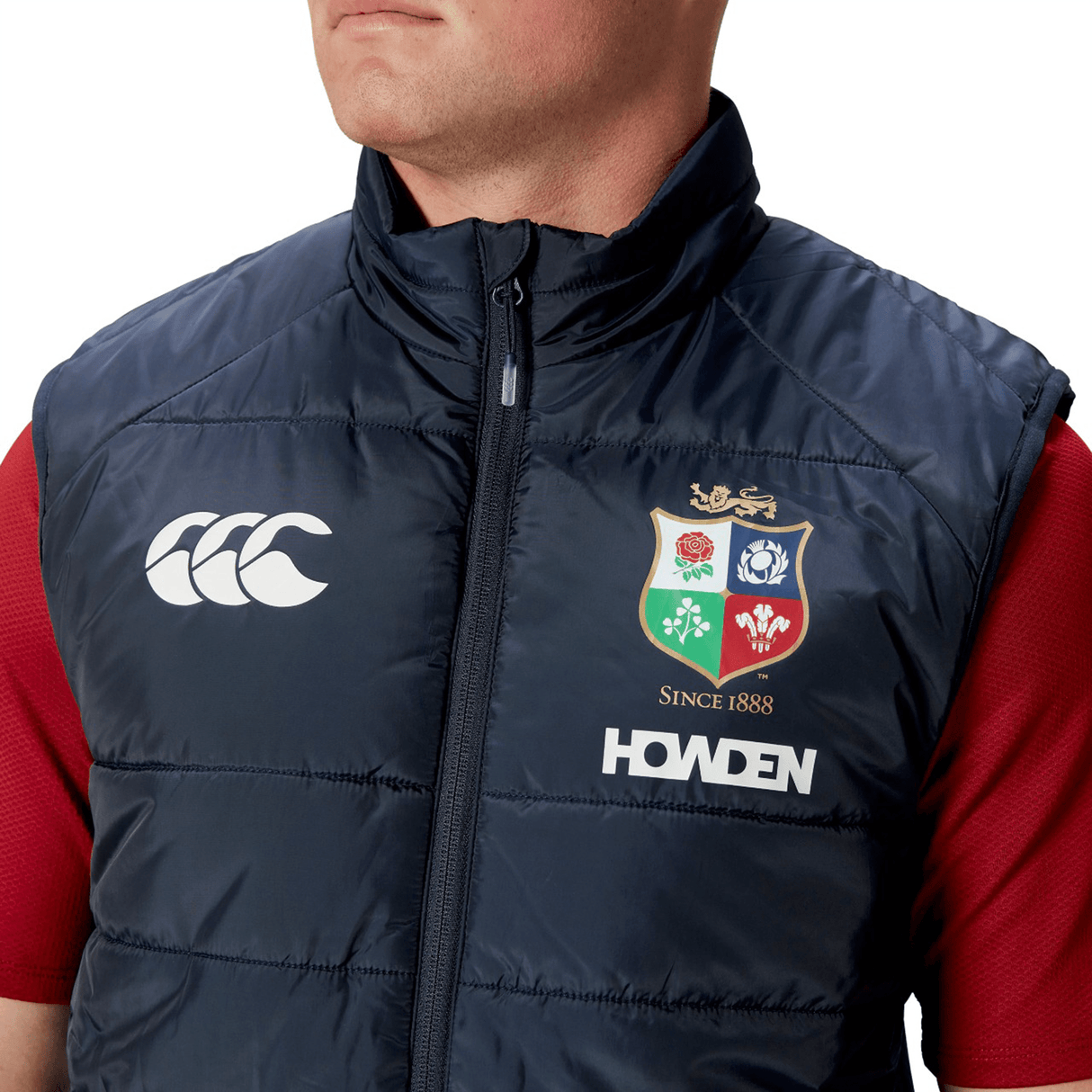 Dressed in a British & Irish Lions 2025 Microlight Gilet by Canterbury, this individual showcases a navy blue vest adorned with the iconic Canterbury logo, the British and Irish Lions crest, and "HOWDEN" lettering. Made from recycled polyester with thermoreg technology, the gilet is fashionably paired over a red shirt.