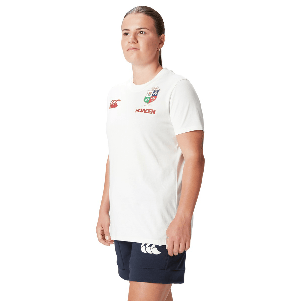 A dedicated rugby supporter stands confidently against a white background, wearing the Women's British & Irish Lions 25 Cotton Jersey Tee by Canterbury, made of organic cotton with logos, and paired with classic black shorts.