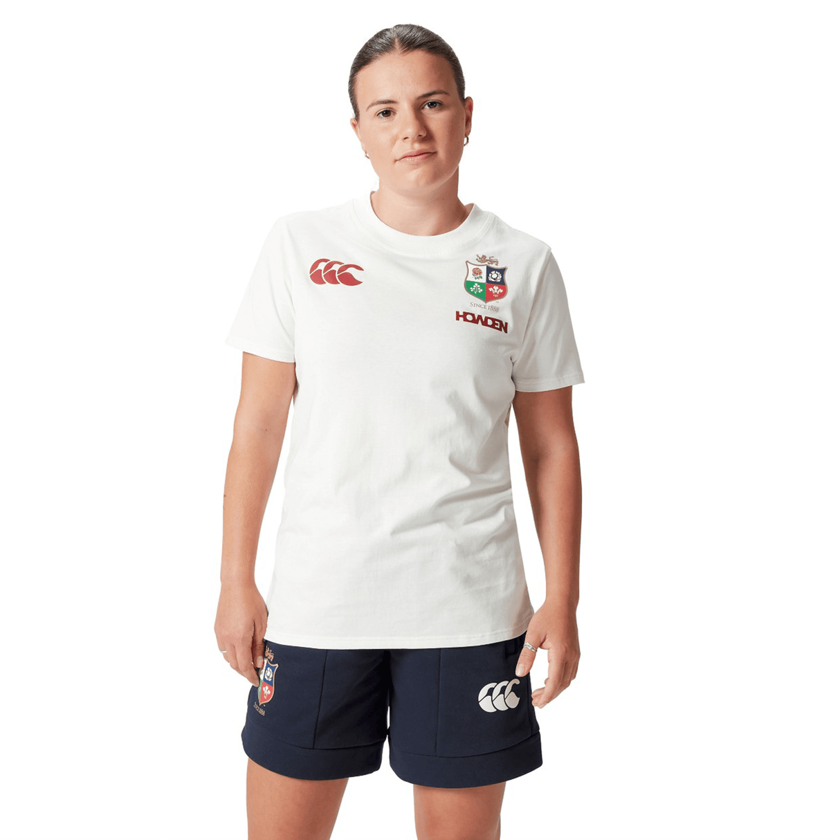A rugby supporter proudly wearing the Women's British & Irish Lions 25 Cotton Jersey Tee by Canterbury, featuring red British & Irish Lions logos on a white background, and dark shorts stands against a white backdrop.