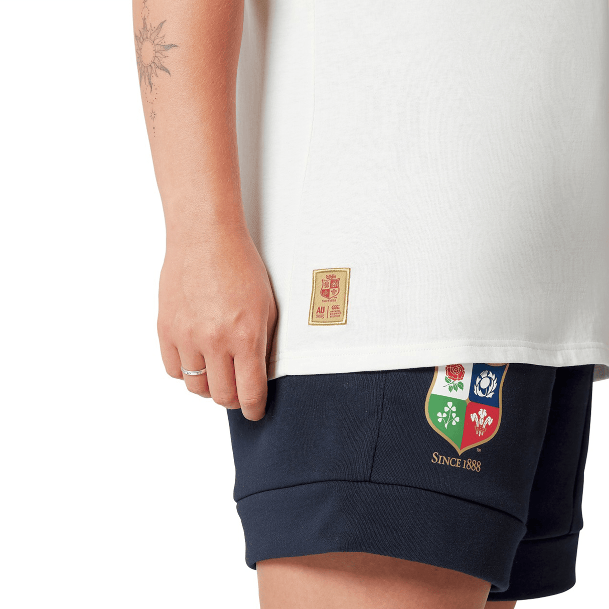 A rugby fan wearing the Women's British & Irish Lions 25 Cotton Jersey Tee by Canterbury, paired with navy shorts adorned with vibrant embroidered patches, stands against a crisp white background. This outfit from Canterbury, crafted from organic cotton, effortlessly combines style and sustainability.