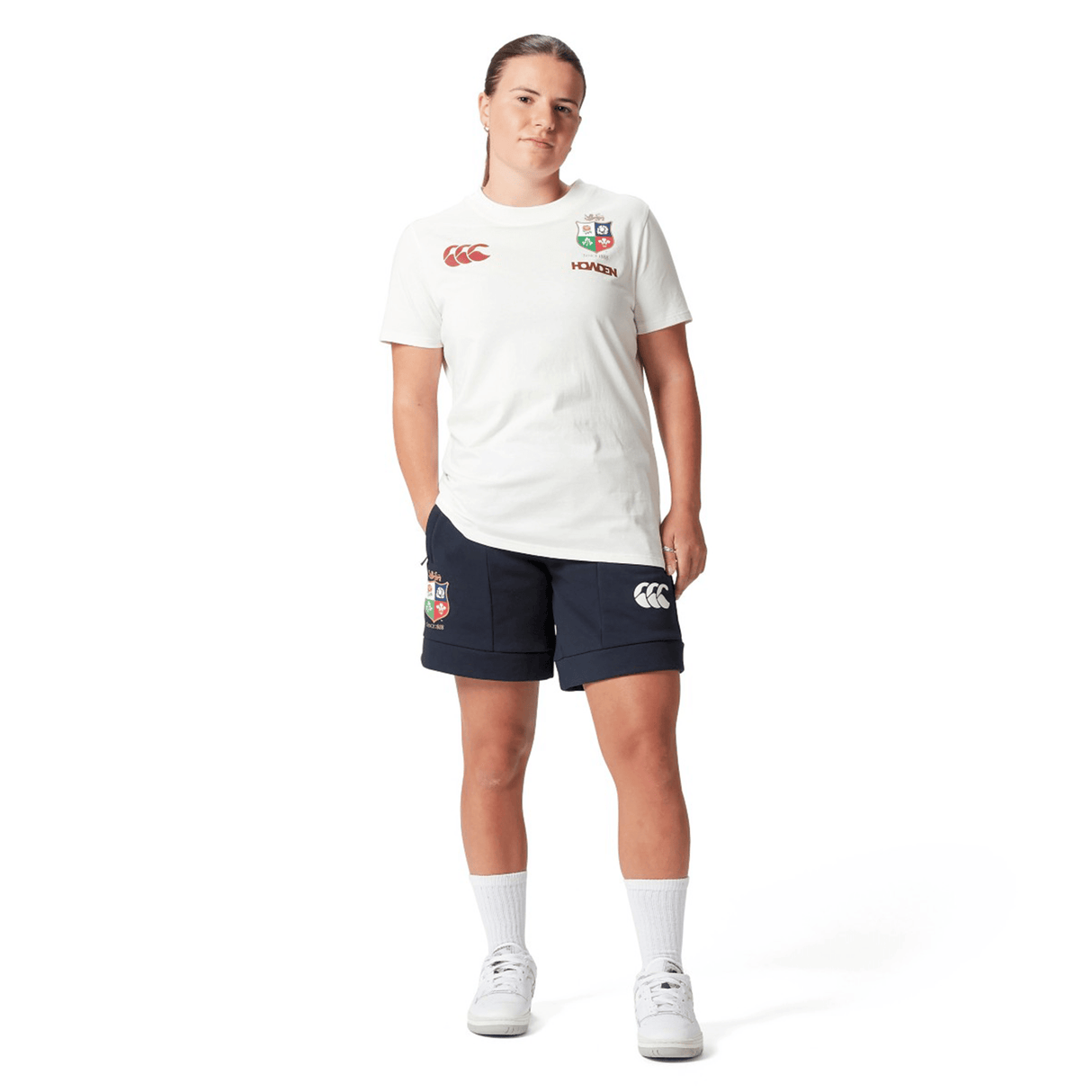 A rugby supporter wears a Women's British & Irish Lions 25 Cotton Jersey Tee by Canterbury, paired with navy shorts featuring logos, complemented by white socks and sneakers against a plain white background. This outfit embodies the essence of the British & Irish Lions.