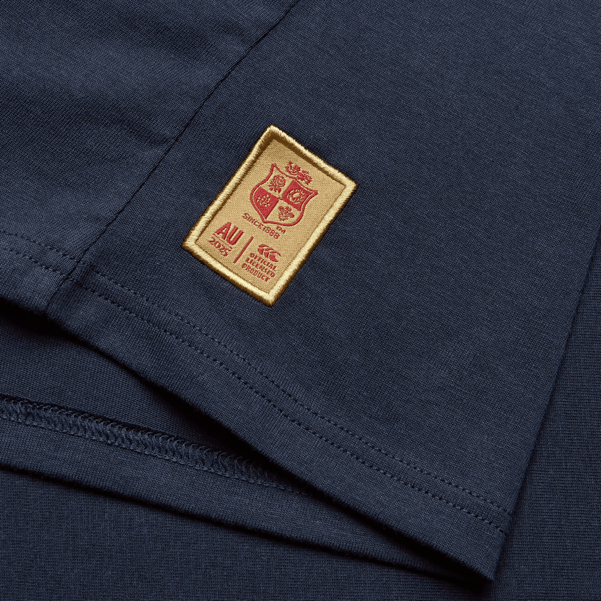 A Women's British & Irish Lions 25 Cotton Jersey Tee by Canterbury, in navy blue, is crafted from organic cotton and features a gold and red stitched label adorned with a crest, the text "AU 2025," along with additional small details underneath, making it perfect for any rugby supporter or fan of the British & Irish Lions.