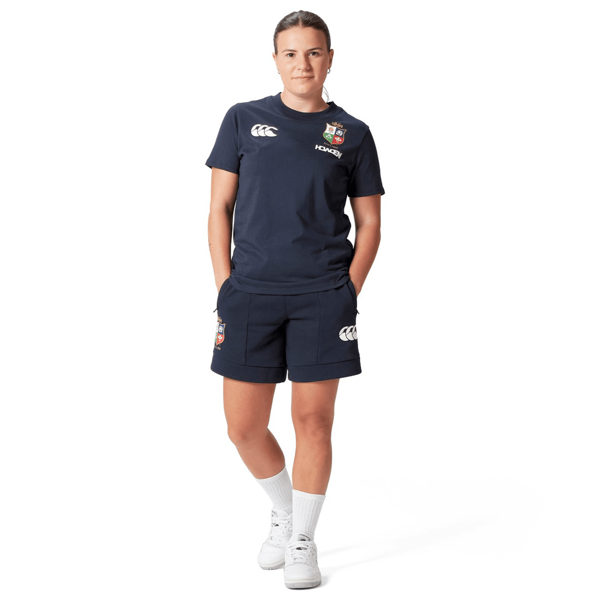A rugby supporter stands confidently in the Women's British & Irish Lions 25 Cotton Jersey Tee by Canterbury, adorned with logo patches. Complemented by white socks and shoes, this ensemble reflects their allegiance. The clean, white background highlights the sporty flair and dedication of a true British & Irish Lions fan.