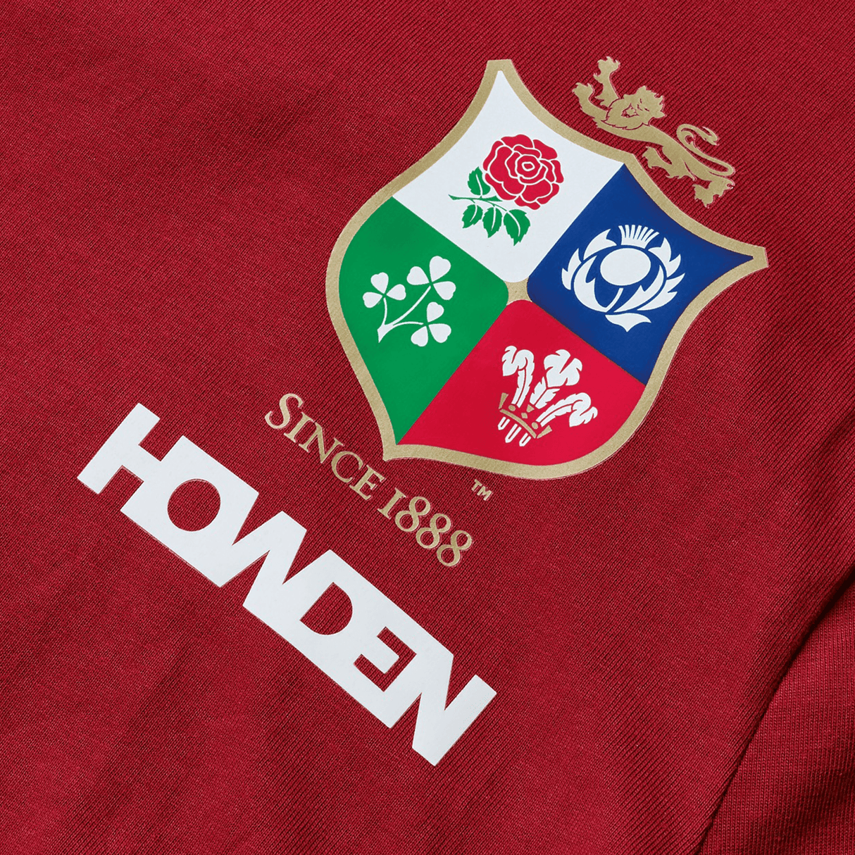 The Youth British & Irish Lions 2025 Cotton Jersey Tee by Canterbury features red fabric with a crest bearing a rose, thistle, shamrock, and feathers, along with "Since 1888" above "HOWDEN." An ideal tribute for the passionate rugby supporter.