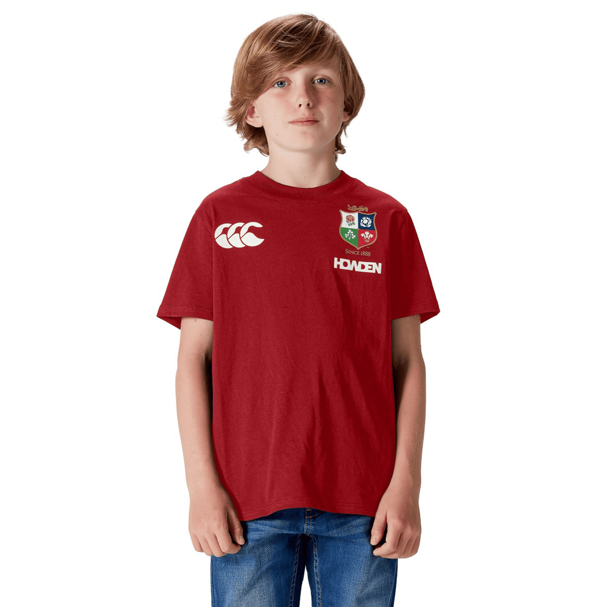 A rugby fan proudly poses against a white backdrop, wearing a Youth British & Irish Lions 2025 Cotton Jersey Tee by Canterbury in red and pairing it with blue jeans.