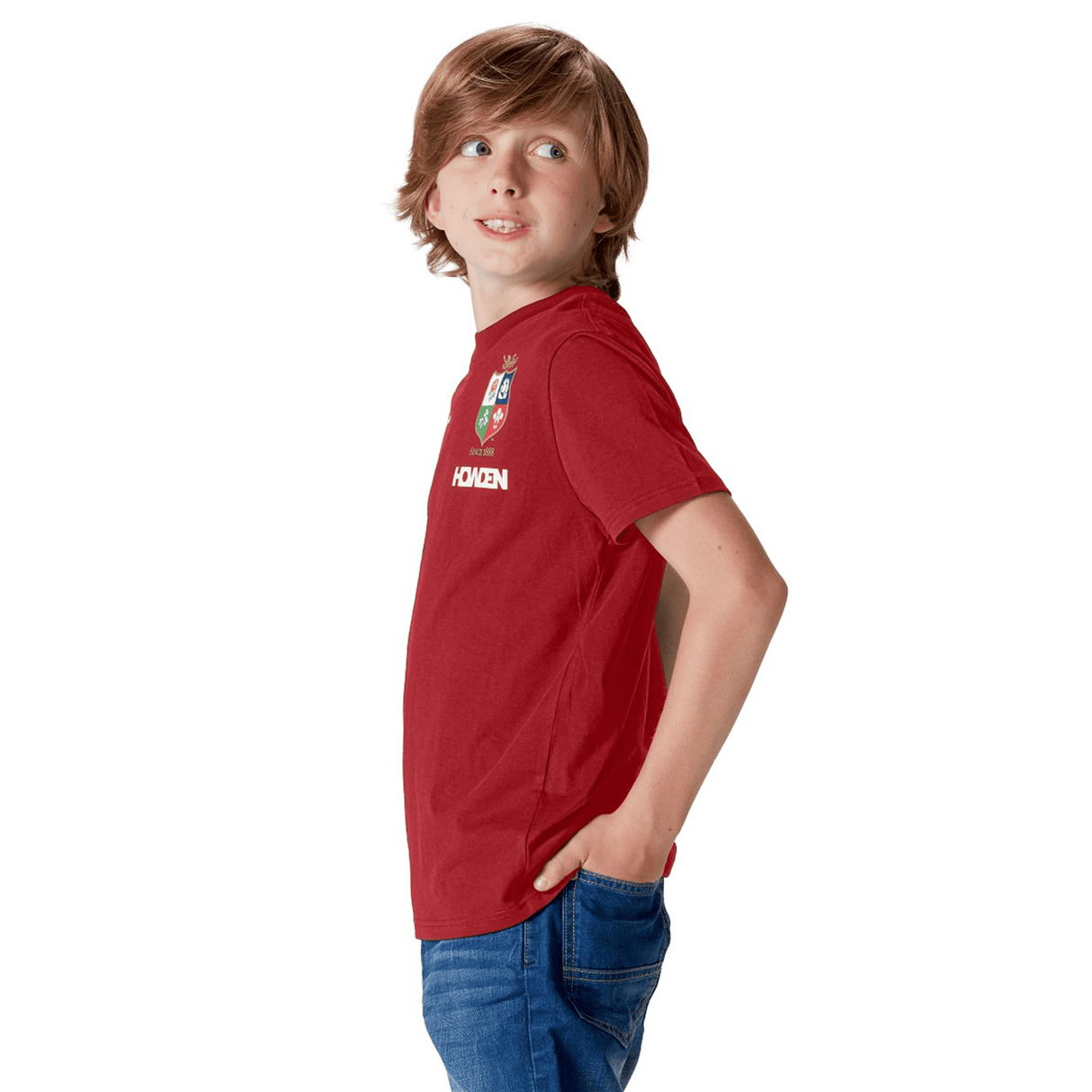 A young rugby fan wearing the Youth British & Irish Lions 2025 Cotton Jersey Tee by Canterbury stands casually with hands in his jeans pockets, glancing to the side against a white backdrop.