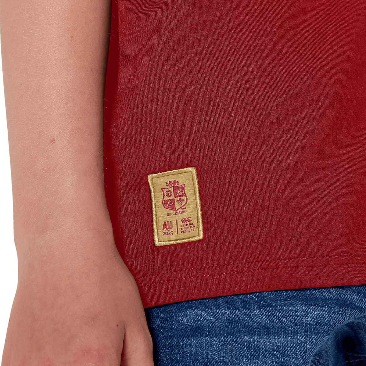 A close-up shows a rugby fan in the Youth British & Irish Lions 2025 Cotton Jersey Tee by Canterbury, made of organic cotton. It features a beige "AU 2025" logo patch. Paired with blue jeans, their hand casually rests near the shirt hem.