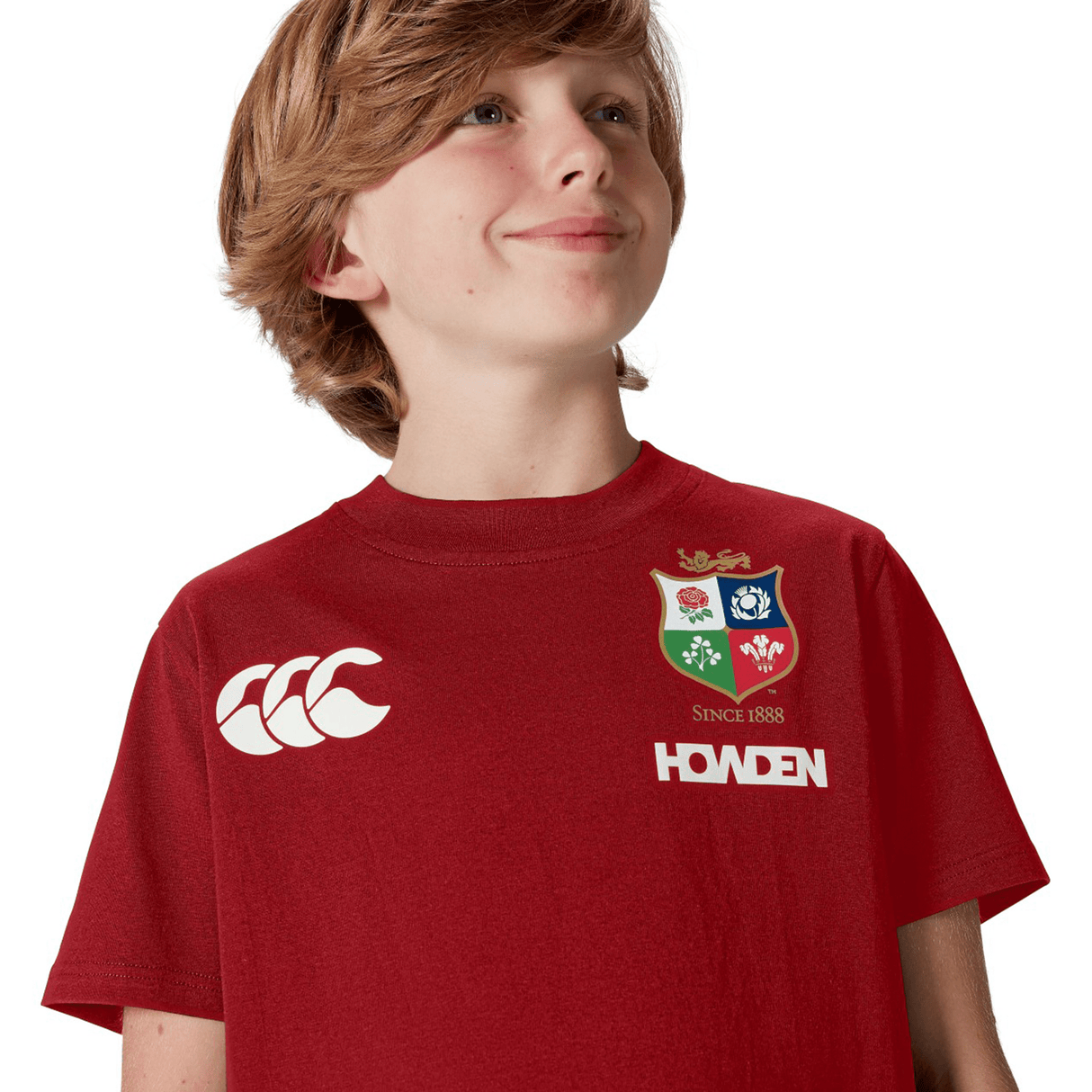 A young rugby fan, wearing a Canterbury Youth British & Irish Lions 2025 Cotton Jersey Tee in red with a logo and text, gazes upwards with a slight smile, capturing the Lion's spirit.
