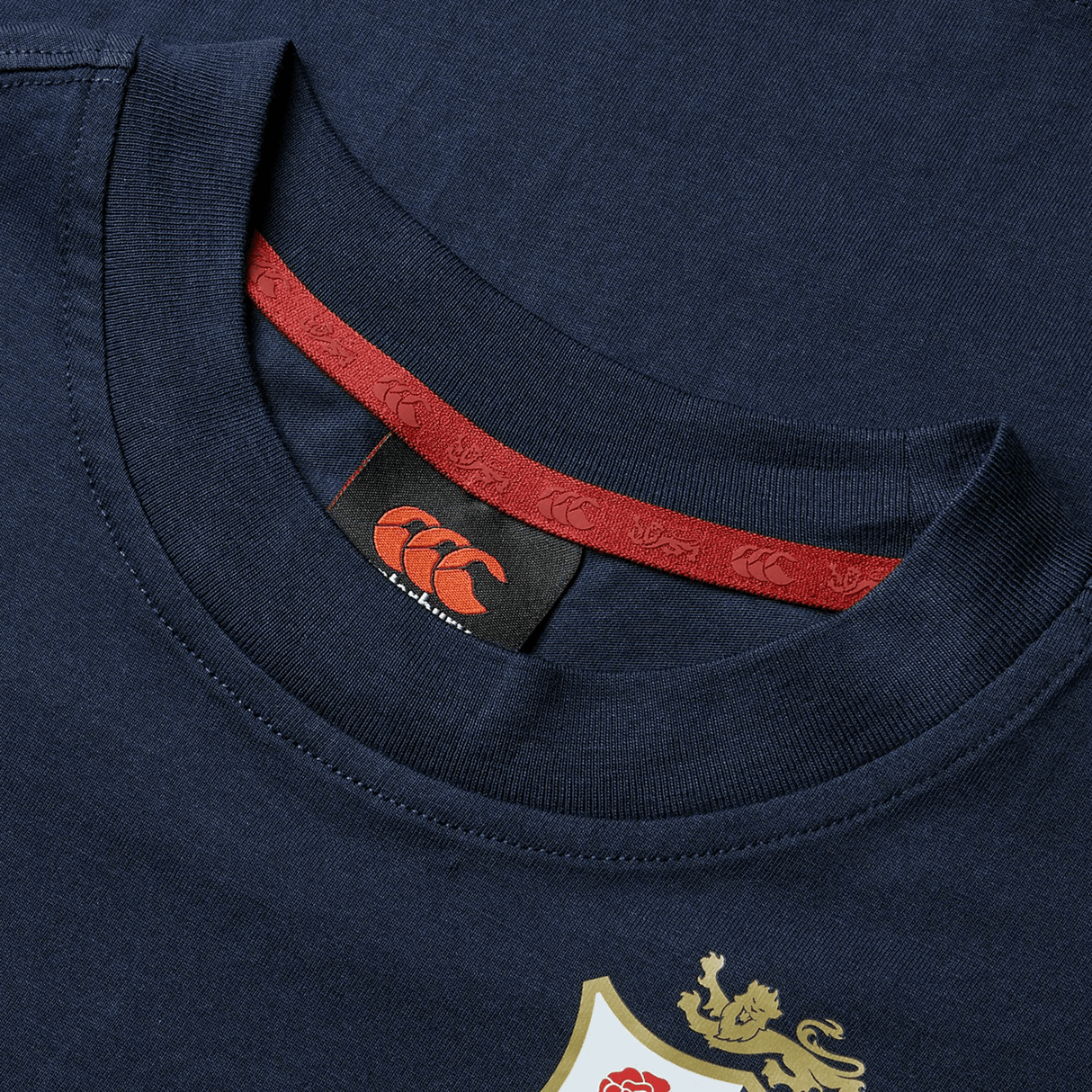 The Youth British & Irish Lions 2025 Cotton Jersey Tee by Canterbury is a navy blue shirt with a red collar band, crafted from organic cotton. It has an orange logo inside and features an emblem with a lion and red rose on the front, perfect for rugby fans cheering on the Lions.