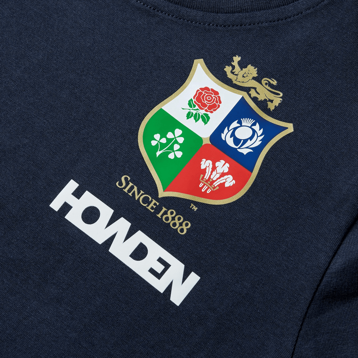 Close-up of the Youth British & Irish Lions 2025 Cotton Jersey Tee by Canterbury, in navy organic cotton. Features a multicolored crest with a lion, rose, shamrock, thistle, and feathers. Ideal for rugby fans with "SINCE 1888" and "HOWDEN" text.