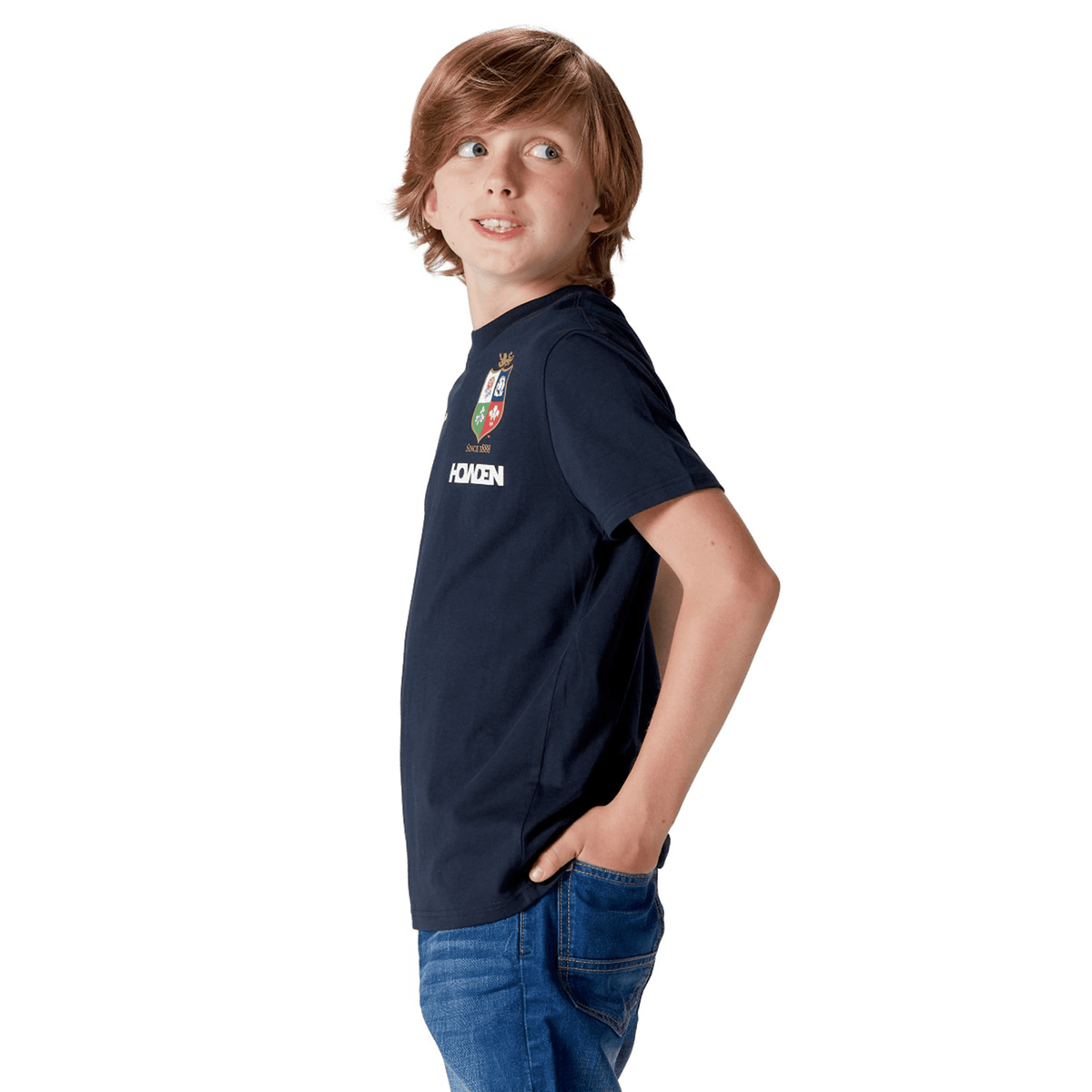 A young rugby fan in a navy Youth British & Irish Lions 2025 Cotton Jersey Tee by Canterbury stands with hands in his jeans pockets, gazing sideways against a plain white background. His jersey, crafted from organic cotton, highlights his commitment to sustainability and the Lions.