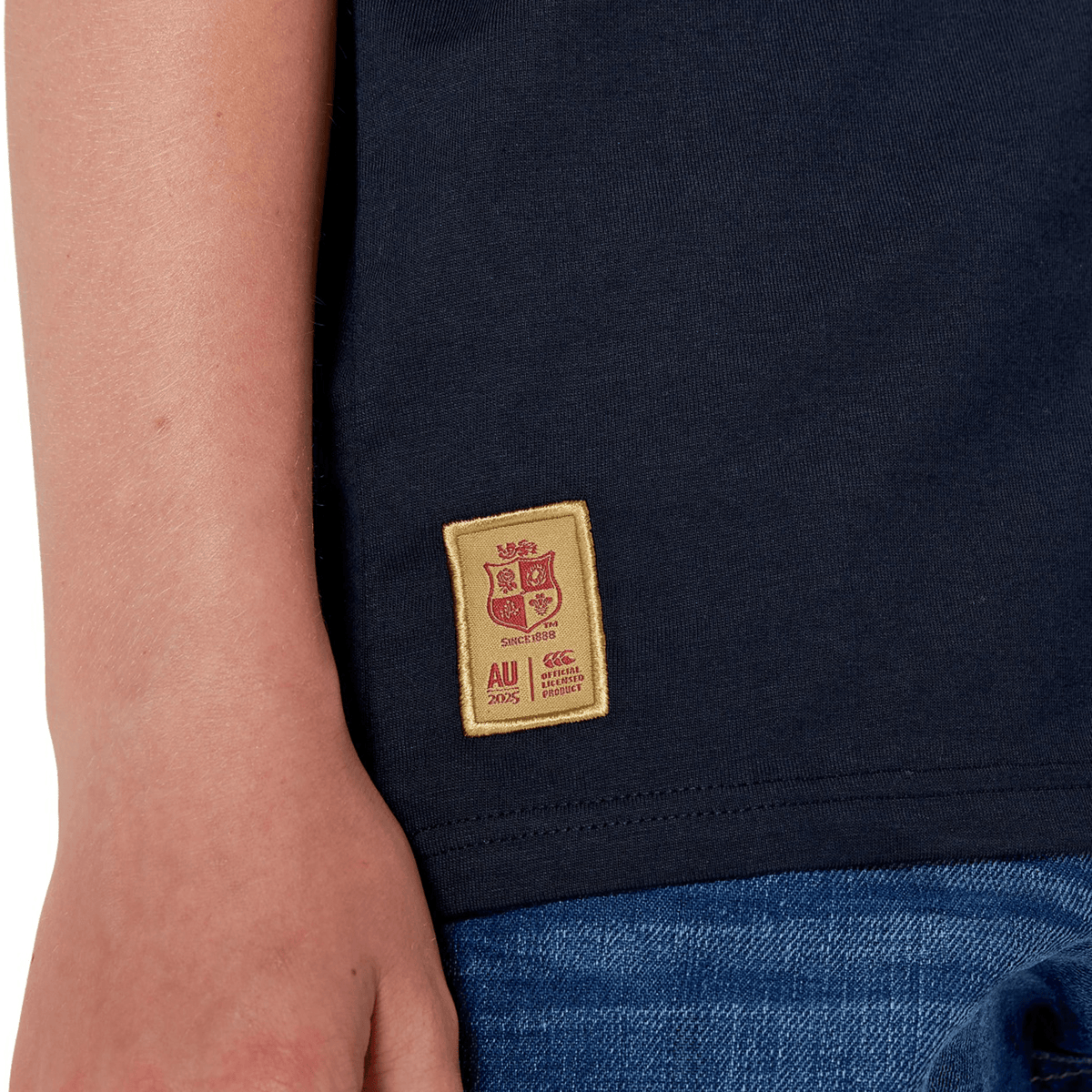 A rugby supporter wears the Youth British & Irish Lions 2025 Cotton Jersey Tee by Canterbury, featuring a stitched crest and "AU 2025" patch, made from organic cotton. Paired with blue jeans, it subtly honors the British & Irish Lions.