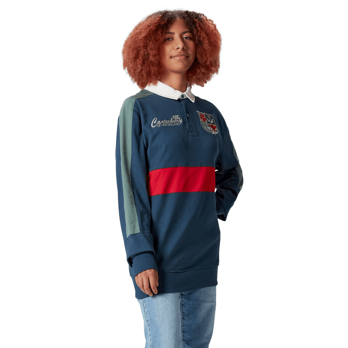 A person with curly hair poses confidently, wearing the British & Irish Lions 2025 Heritage Jersey by Canterbury, featuring a navy design with a red stripe and emblem, embodying the classic Canterbury style, one hand tucked casually in their pocket.
