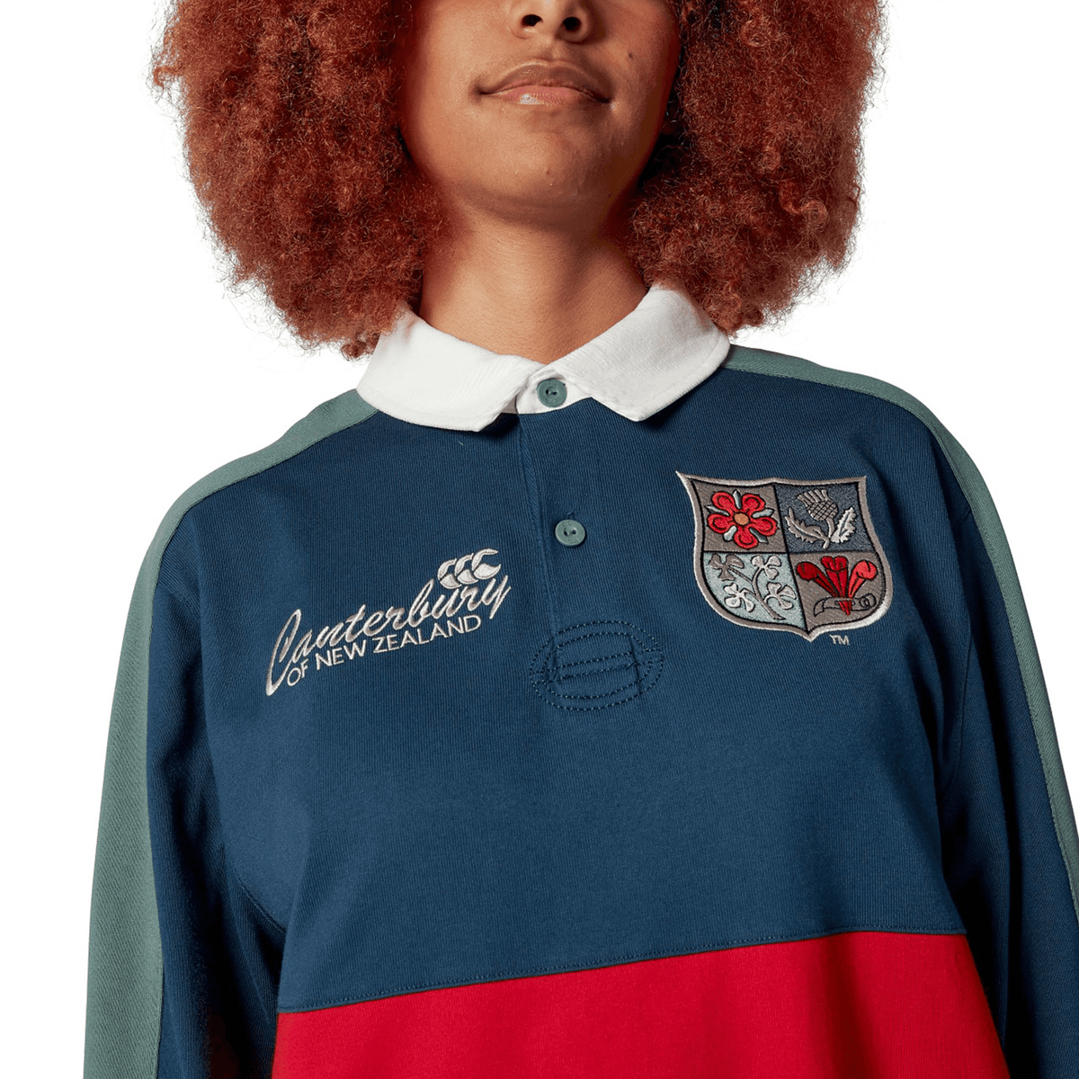 Sporting the British & Irish Lions 2025 Heritage Jersey by Canterbury, a person wears a rugby-style shirt adorned with a logo and crest. It is distinguished by its white collar and vibrant red, blue, and green color blocks, echoing the classic Canterbury Heritage design.