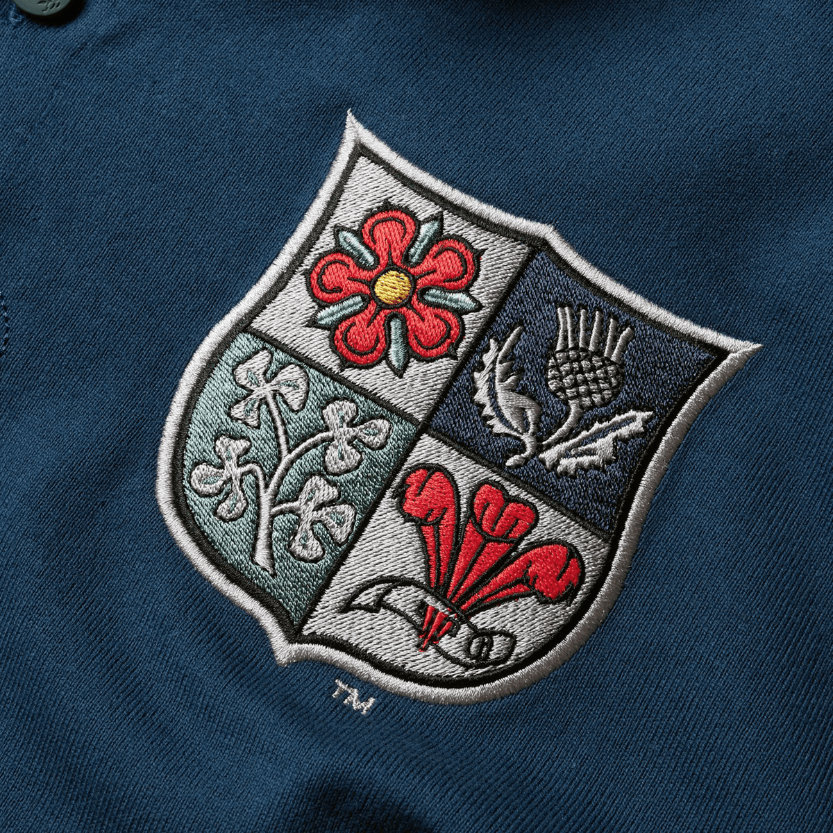 The British & Irish Lions 2025 Heritage Jersey by Canterbury features an embroidered emblem on blue fabric, showcasing a red flower, a thistle, shamrocks, and a red dragon over green, white, and blue quadrants.