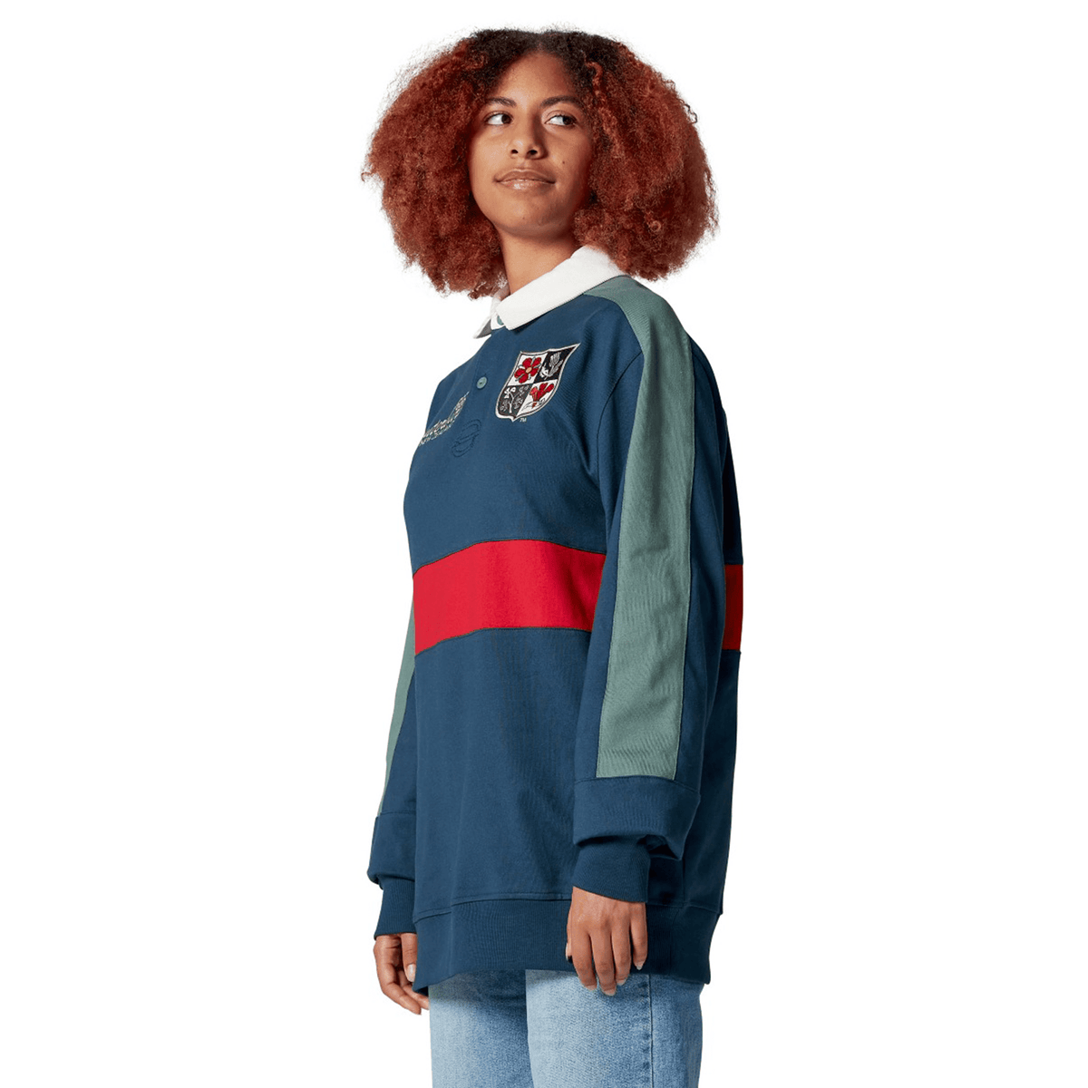 A woman with curly hair wears the British & Irish Lions 2025 Heritage Jersey by Canterbury, showcasing a blue and green design with a red stripe and logo, paired with jeans. She gazes thoughtfully to the side.