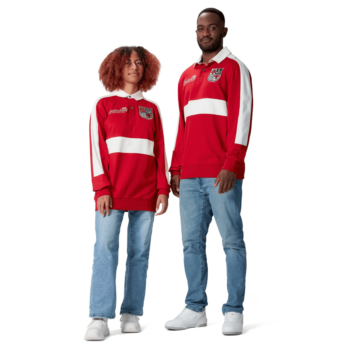 Two individuals wearing coordinated red and white long-sleeve British & Irish Lions 2025 Heritage Jerseys by Canterbury, paired with jeans, stand against a white background.