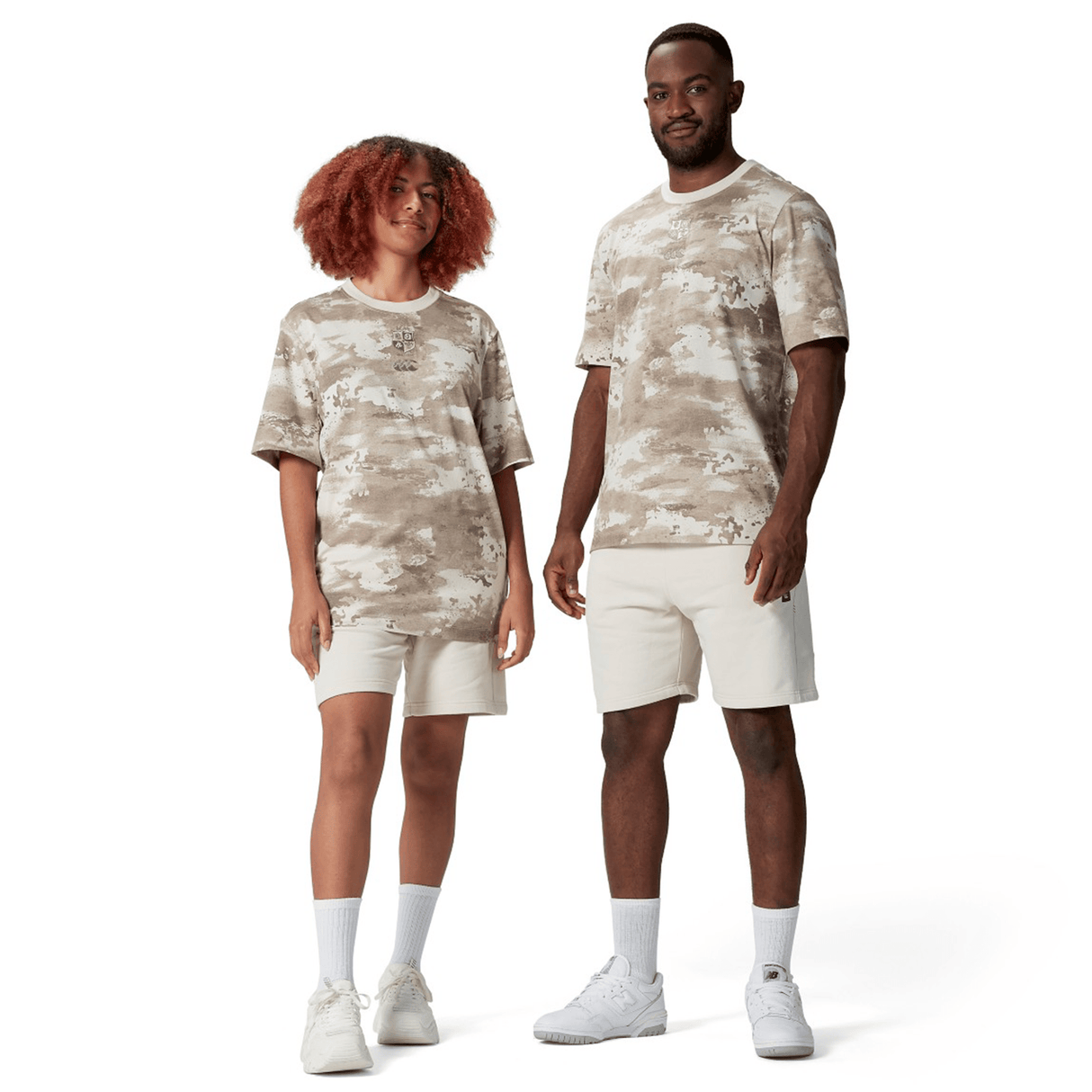 Two individuals stand on a plain white background, each wearing the British & Irish Lions 2025 Camo Tee by Canterbury, paired with beige desert taupe shorts and crisp white sneakers.