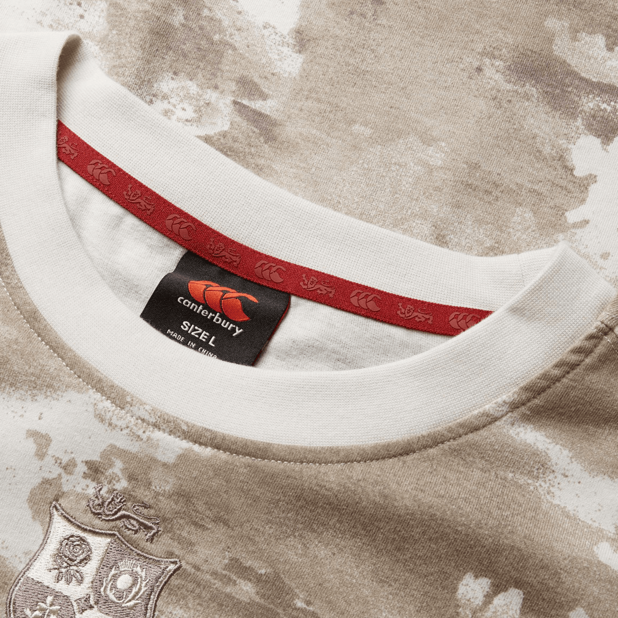 A detailed view of the British & Irish Lions 2025 Camo Tee by Canterbury shows a Desert Taupe and white pattern, complete with a red tag displaying the Canterbury logo and size "L." The fabric is adorned with an elegant crest design, evocative of the British & Irish Lions.