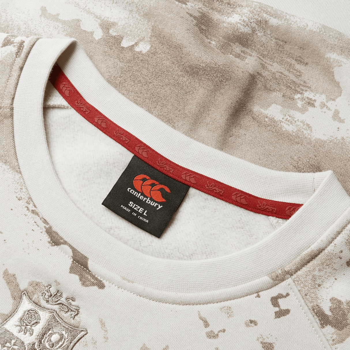 Close-up of a beige and white camouflage patterned sports jersey from Canterbury, reminiscent of the British & Irish Lions 25 Camo Crew Sweatshirt. It includes a red inner collar with logo design and a visible "2XL" size label on the neck tag, reflecting the essence of British & Irish Lions gear.