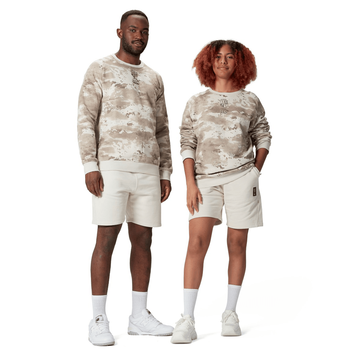 Two individuals pose side by side, donning identical beige British & Irish Lions 25 Camo Crew Sweatshirts by Canterbury, paired with shorts. Both are wearing white sneakers and socks, embodying a relaxed and casual style. Their outfits reflect the stylish comfort associated with Canterbury, renowned for its British & Irish Lions collections.