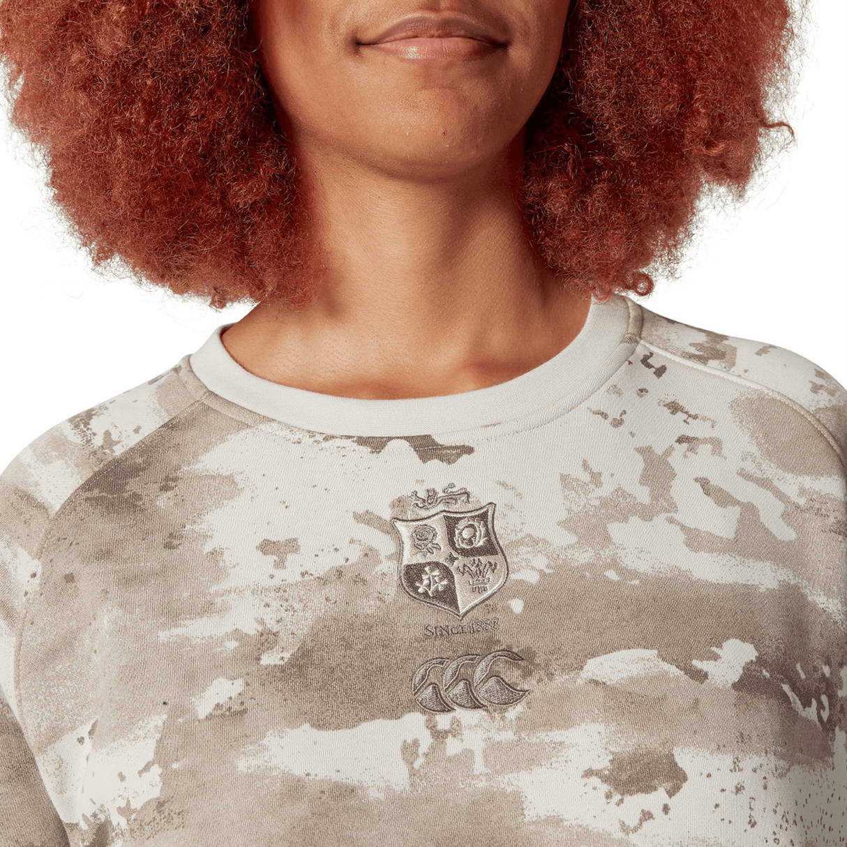 A person with curly, reddish hair is wearing the British & Irish Lions 25 Camo Crew Sweatshirt by Canterbury, featuring a crest and logo.