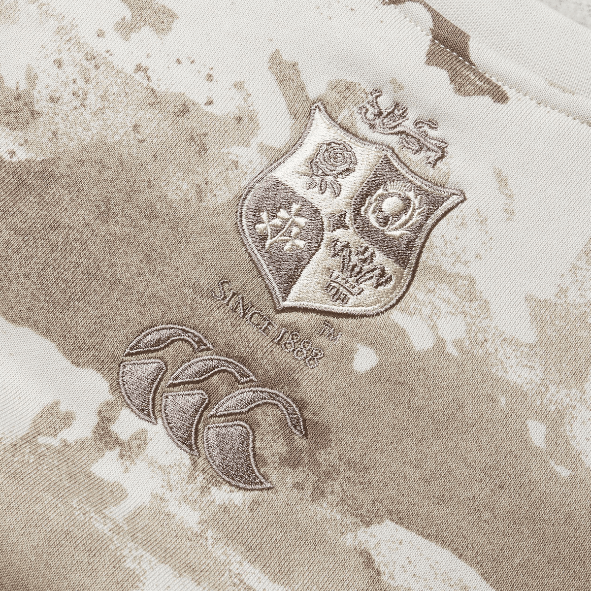 A close-up of the British & Irish Lions 25 Camo Crew Sweatshirt by Canterbury showcases a beige and brown rugby jersey design. The piece proudly displays the prestigious British & Irish Lions crest, accompanied by the text "Since 1888" and three stylized logos underneath, exemplifying Canterbury's timeless craftsmanship.