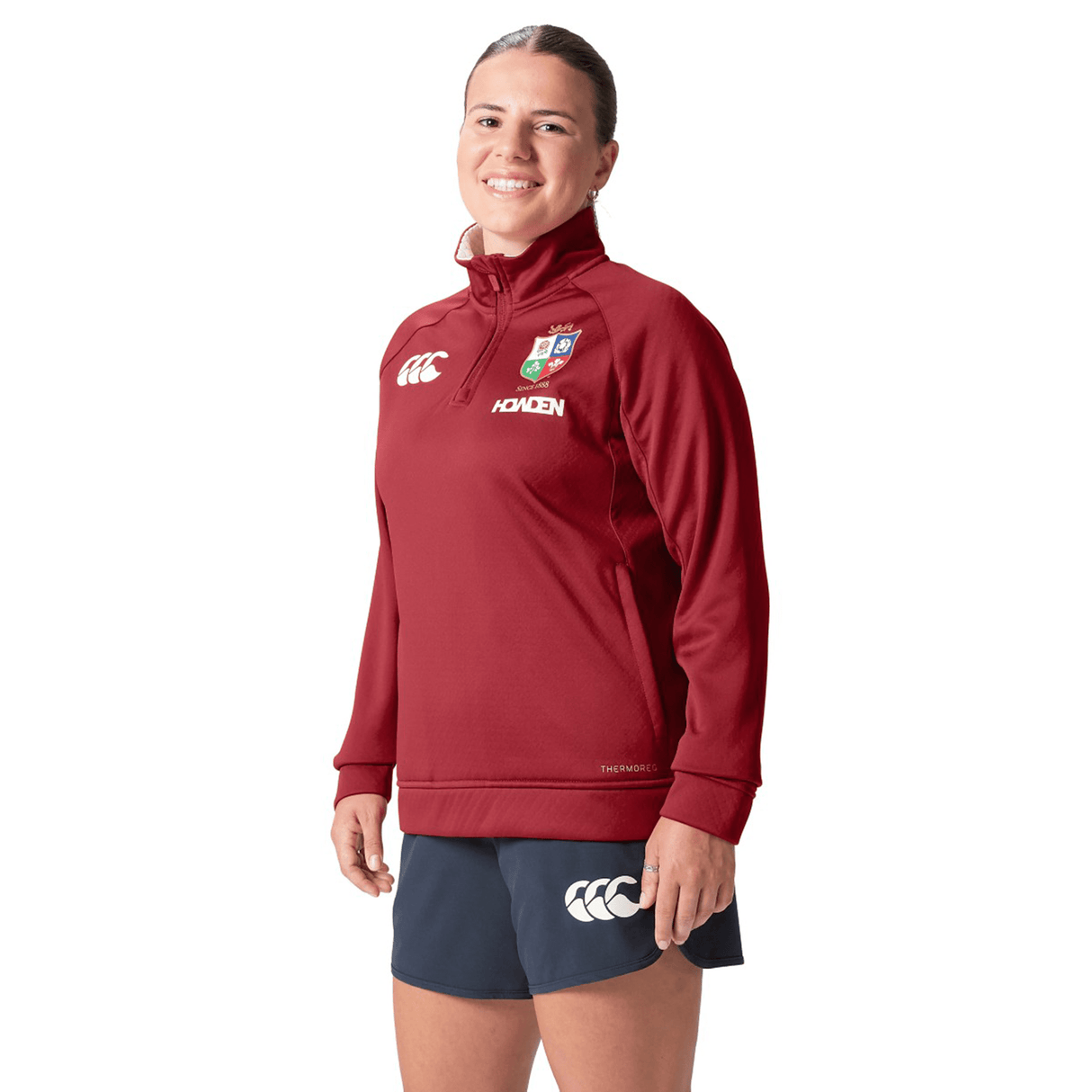 A person is wearing the Women's British & Irish Lions 2025 1/4 Zip Fleece by Canterbury, paired with navy shorts, standing and smiling against a plain background.