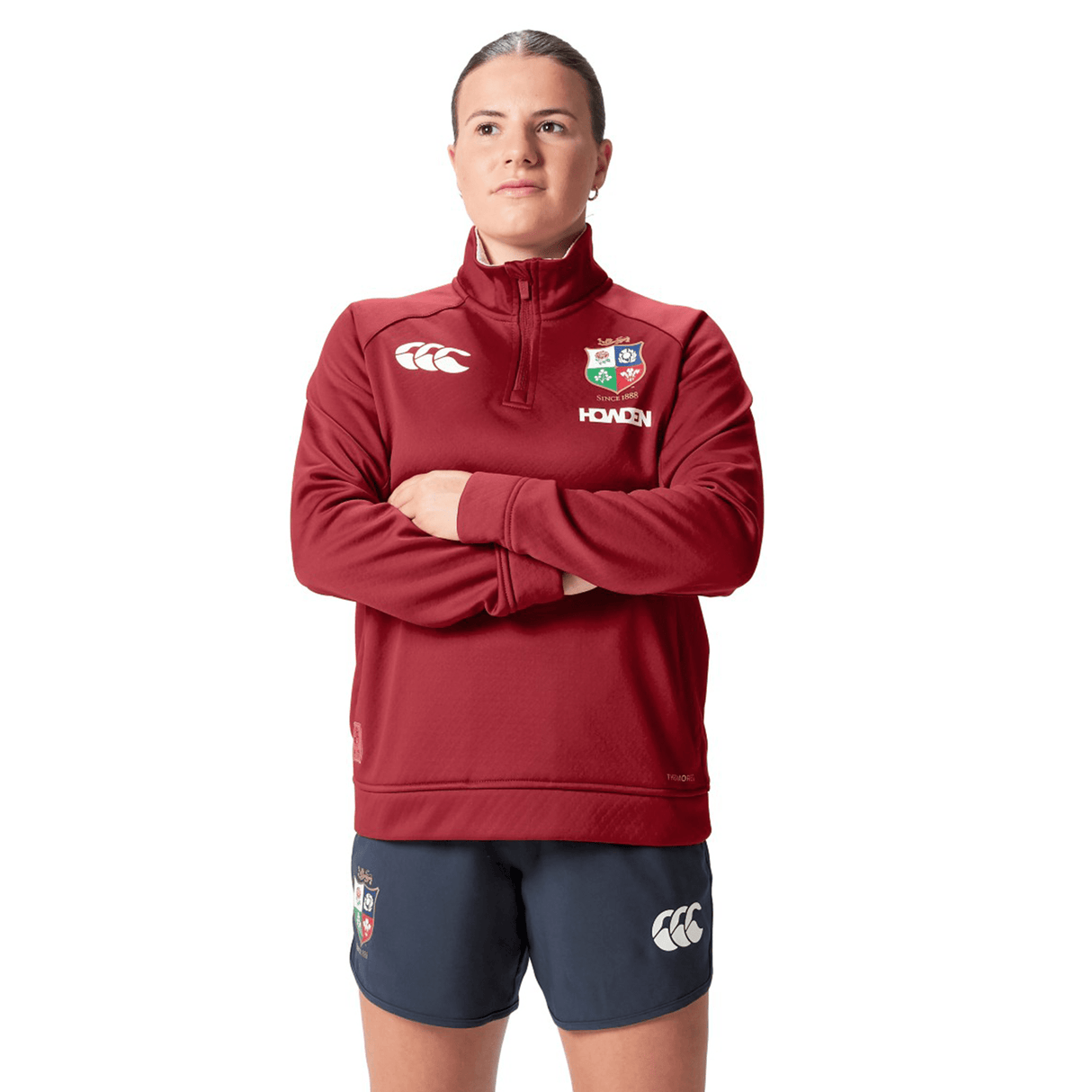 A person wearing a Women's British & Irish Lions 2025 1/4 Zip Fleece by Canterbury, which has a maroon design reminiscent of their style, along with navy shorts, stands with arms crossed. The jacket showcases a white logo and a colorful emblem in its women's fit style.