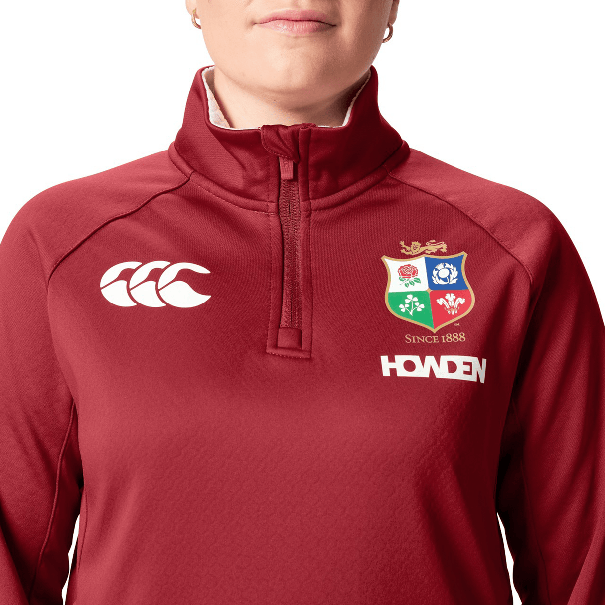 Dressed in a red Women's British & Irish Lions 2025 1/4 Zip Fleece by Canterbury, adorned with logos including a shield emblem and the text "HOWDEN," a person stands confidently, highlighting the sleek Women's Fit style.
