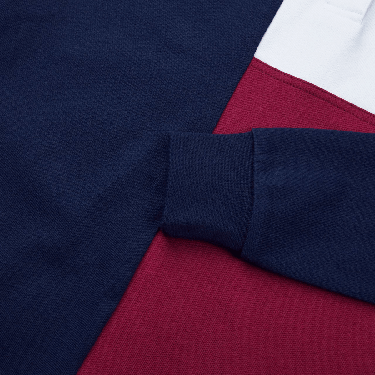 A close-up of the Women's Canterbury Harlequin Weber Long Sleeve Rugby Jersey fabric displays navy, red, and white color blocks. It features a long sleeve with a navy cuff adorned with the Canterbury logo diagonally positioned on the textile.