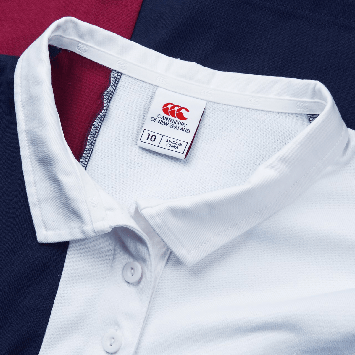 Close-up of the Women's Canterbury Harlequin Weber Long Sleeve Rugby Jersey, a white collared sports jersey showcasing the Canterbury logo, set against a lively maroon and navy fabric backdrop.