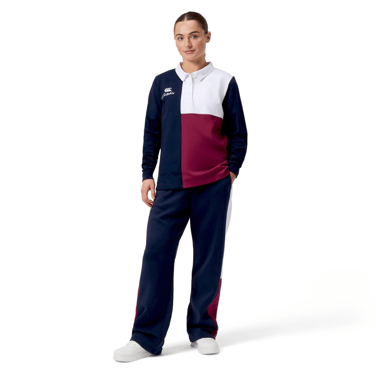 A woman stands against a white backdrop in the Canterbury Women's Harlequin Weber Long Sleeve Rugby Jersey, featuring navy, burgundy, and white blocks. She pairs the iconic harlequin top with matching pants and rests her hands in the pockets.