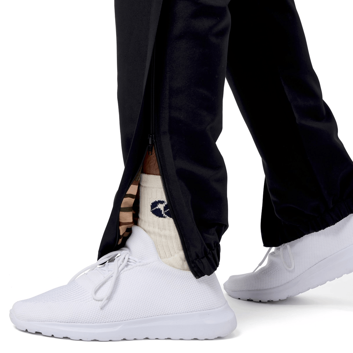Wearing a stylish twist on classic attire, the person pairs white sneakers with white socks featuring a blue logo and Canterbury's Uglies Cuffed Stadium Pant in black. The pants, with zippers partially open at the ankles, deliver a dynamic look suitable for casual outings and athletic activities.