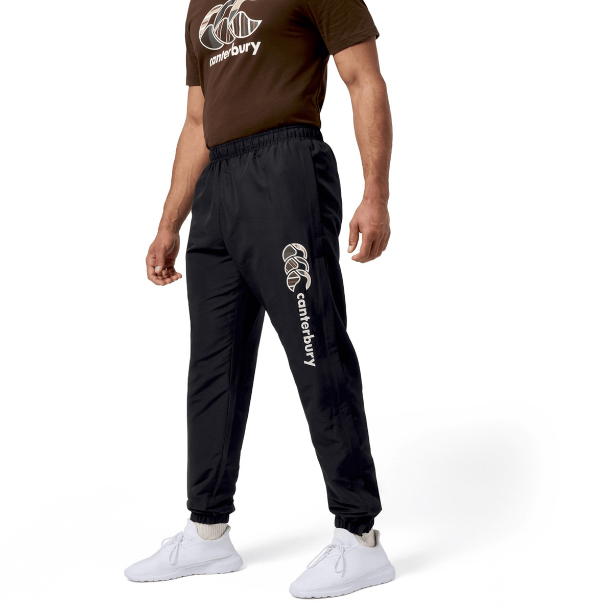 A man stands against a white background, showcasing his relaxed rugby style in a brown Canterbury Uglies T-shirt, black Canterbury Uglies Cuffed Stadium Pant with the logo, and white sneakers.