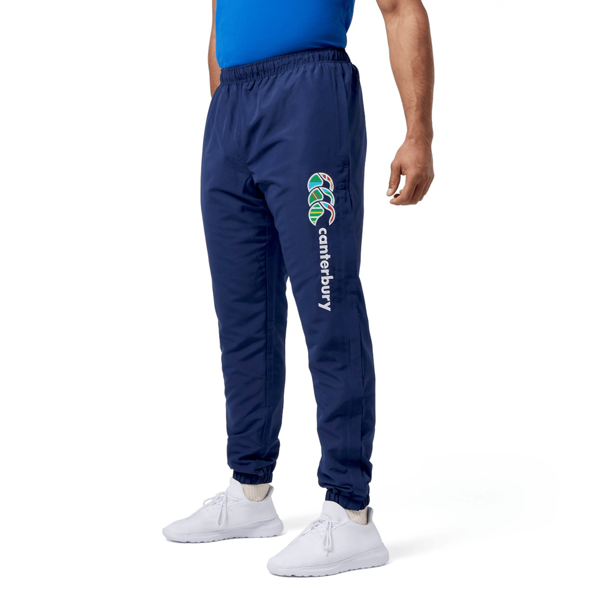 Someone dressed in blue Canterbury Uglies Cuffed Stadium Pants with a vibrant logo and "Canterbury" on the side, paired with a blue top and white sneakers, showcasing a laid-back rugby style.