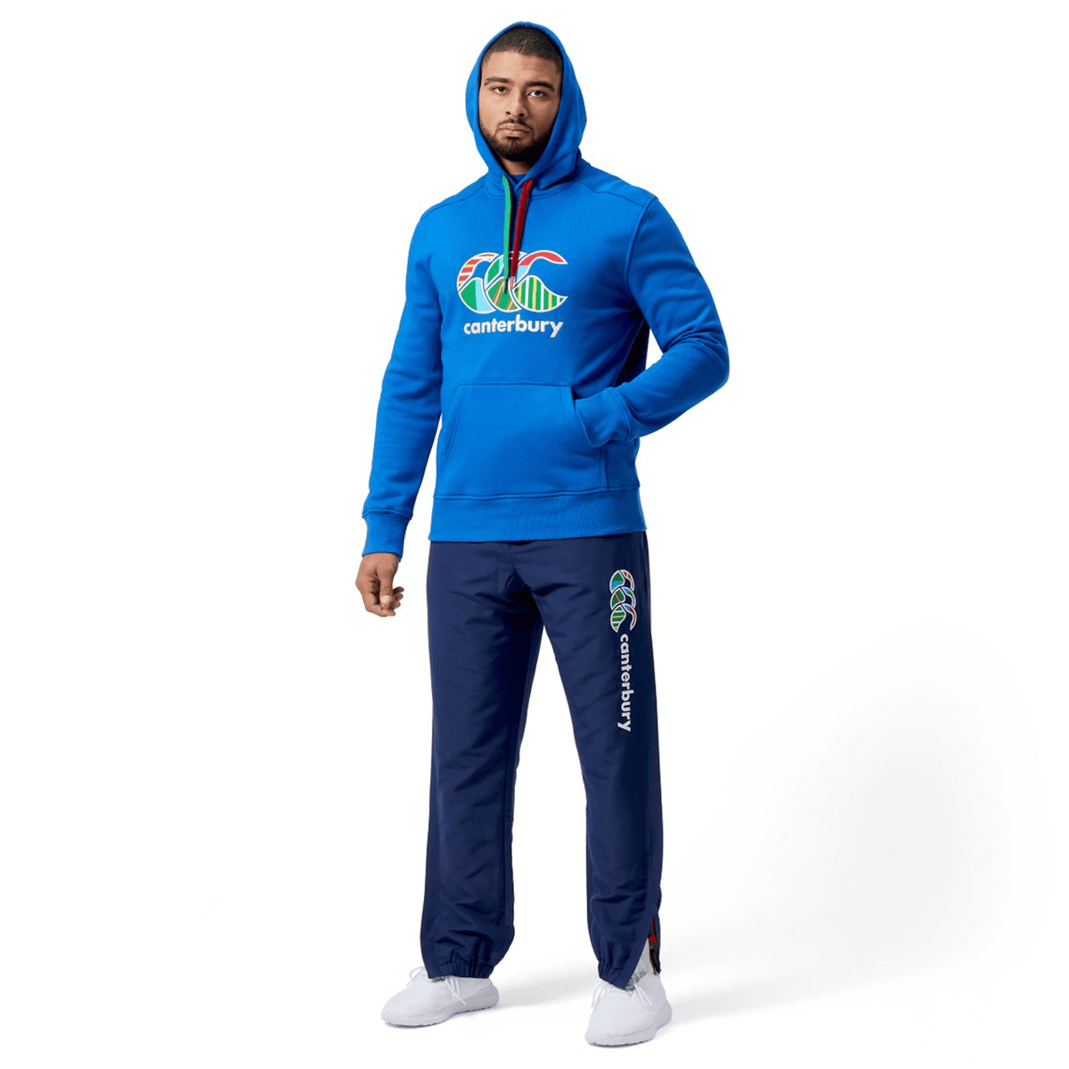 A person in a blue Canterbury Uglies hoodie and Cuffed Stadium Pant, featuring a vibrant rugby-style design, stands against a plain white background showcasing the logo branding.