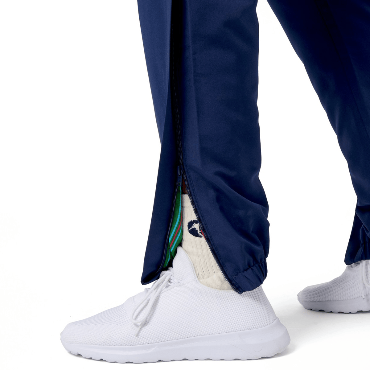 A person showcases their sporty ensemble, wearing white sneakers and green-red striped socks. Complementing the look are Canterbury's blue Uglies Cuffed Stadium Pants, featuring a side zipper reminiscent of Rugby Style, all set against a crisp white background.